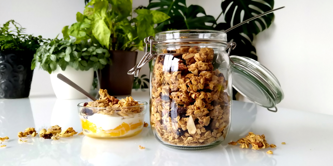 Cashew-sinaasappelgranola met cranberry’s, yoghurt en sinaasappel met een volle pot granola ernaast